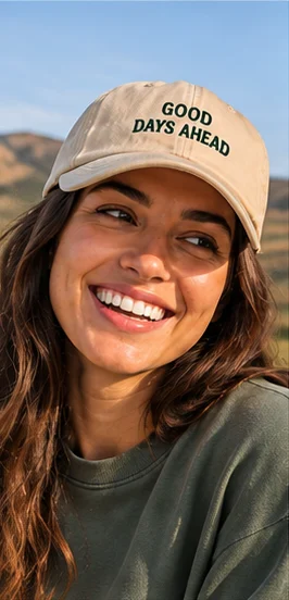 Mulher sorrindo com boné Good Days Ahead bege, montanhas ao fundo
